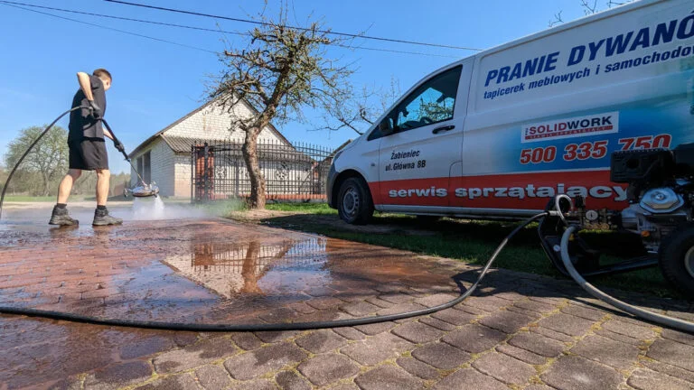Czerwona kostka brukowa silnie zabrudzona w trakcie czyszczenia przez pracownika firmy Solid Work, specjalną maszyną, fragmenty już czyste wyglądają jak nowe. Obok zaparkowana furgonetka firmy oklejona jej reklamą.
