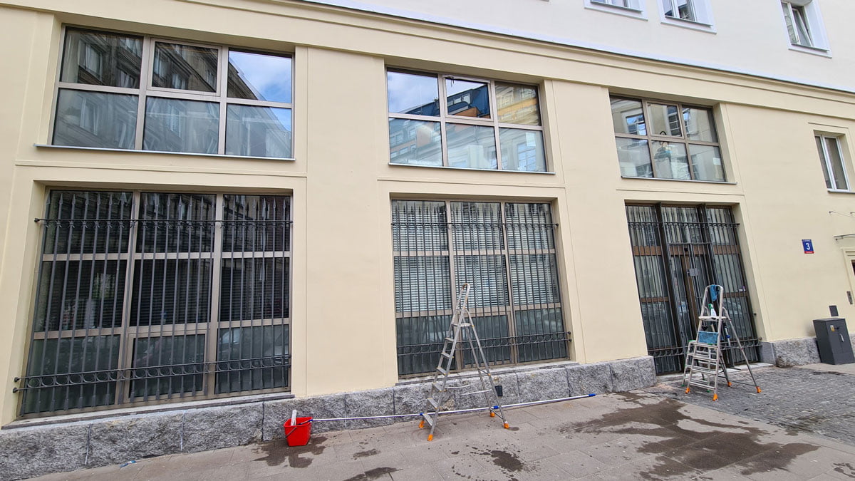 Elewacja budynku z okratowanymi oknami w trakcie ich czyszczenia. Drabiny ustawione przy tych oknach.