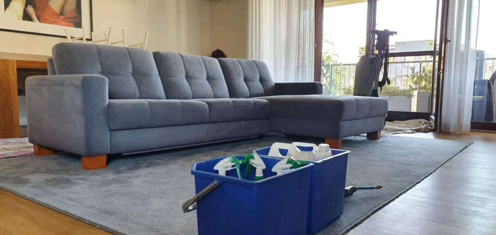 Odnowiona i wyczyszczona duża kanapa narożna na czystym dywanie w przestronnym salonie - mebel oraz dywan w kolorze szaroniebieskim. Przed meblem stoją kubły z preperatami do czyszczenia